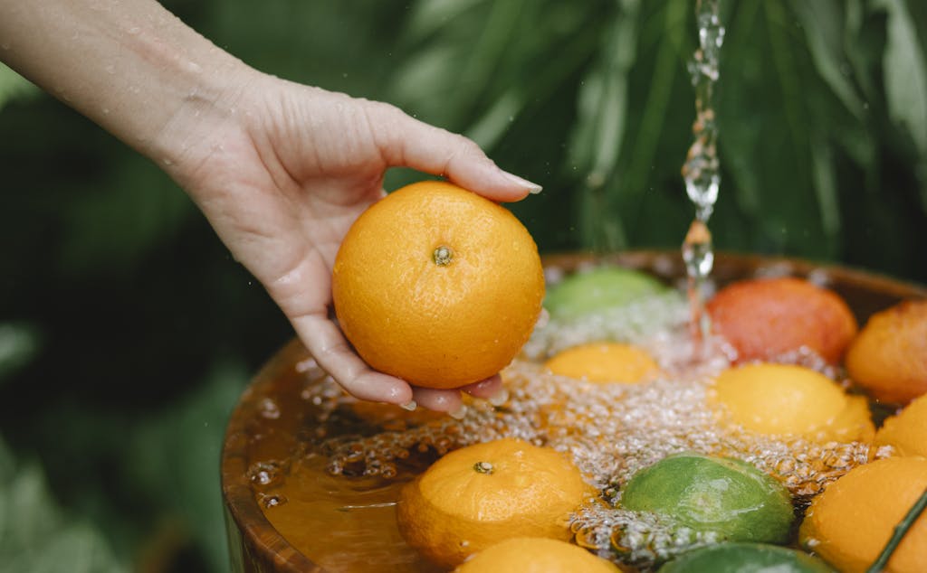 Crop anonymous female washing juicy mandarins under water stream above wooden bowl in lush summer garden